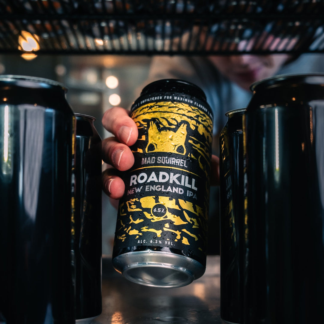 Person holding a can of 'Roadkill' New England IPA between two black mugs in a bar setting.